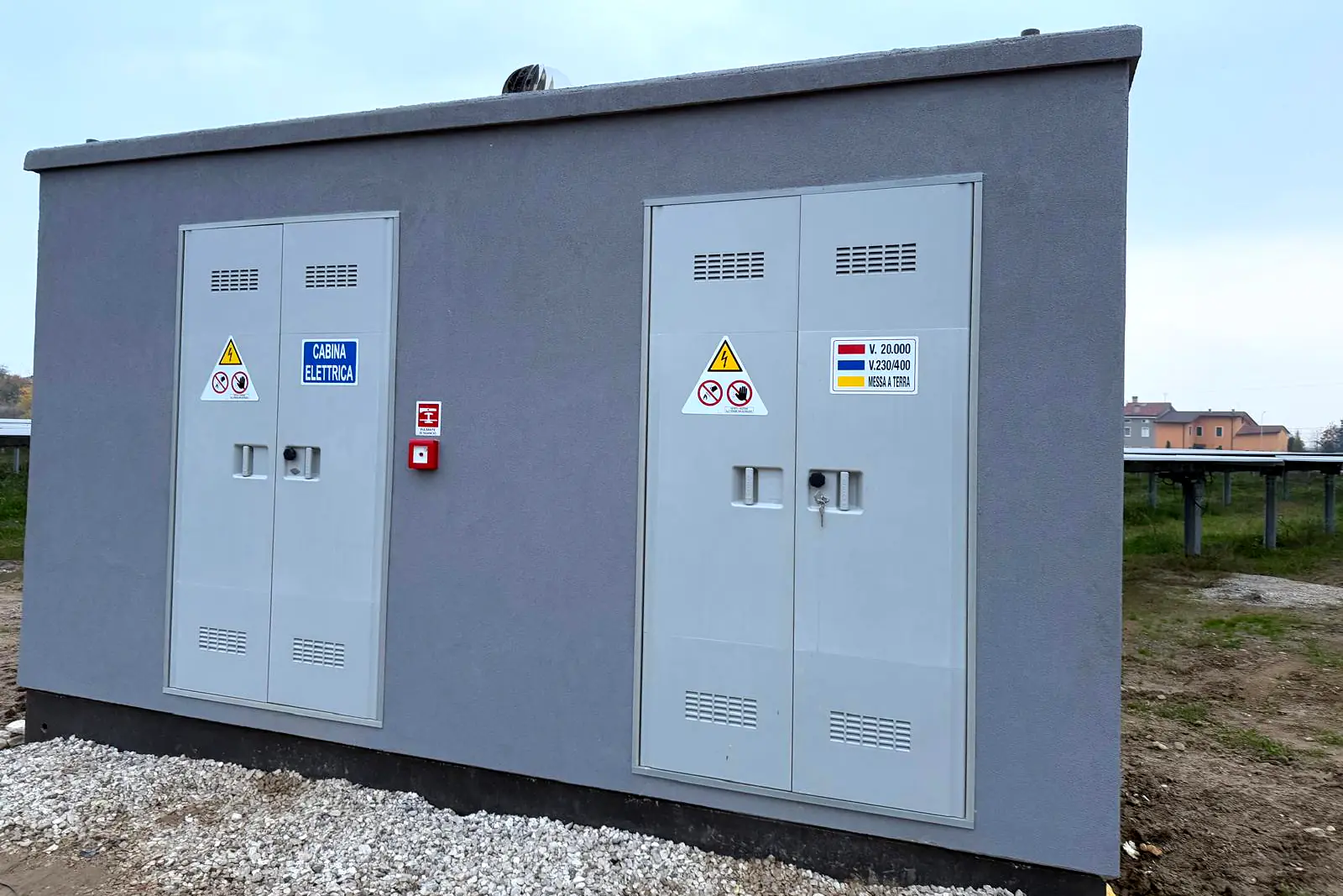Cabina elettrica prefabbricata grigia con due porte doppie, posata accanto a un impianto fotovoltaico